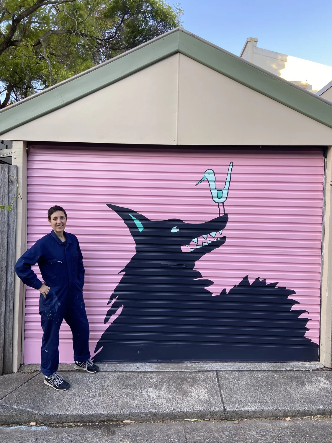 MC in painting overalls beside a mural of a dark dog and a pale bird on a pink roller door