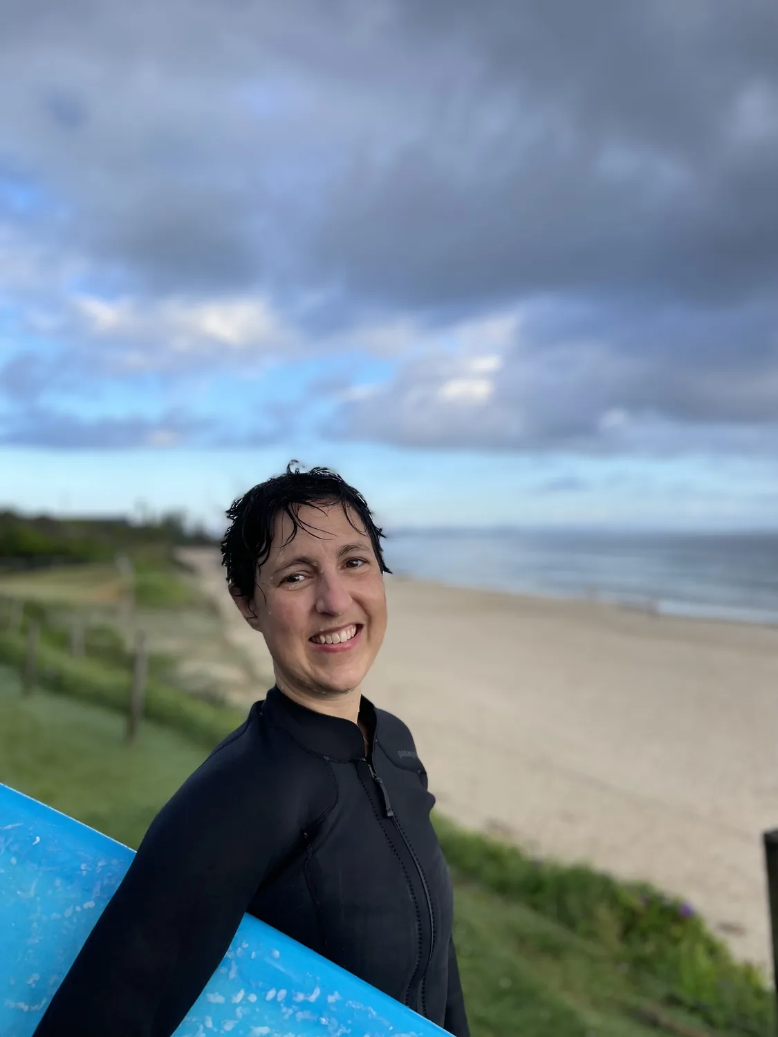 MC in a wetsuit carrying a blue surfboard above the beach